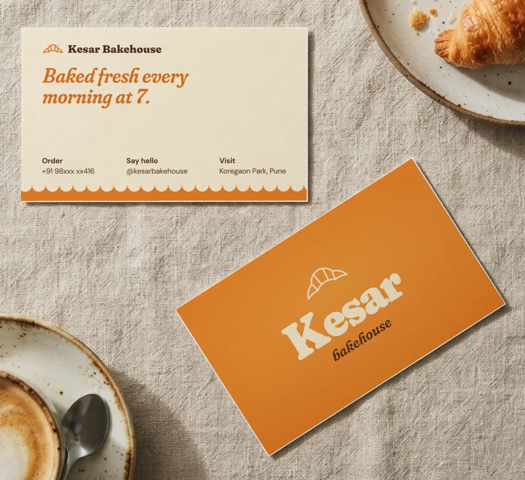 Saffron bakery business cards with a croissant logo, and a cream back with a scalloped edge, on a linen tablecloth beside a croissant and coffee