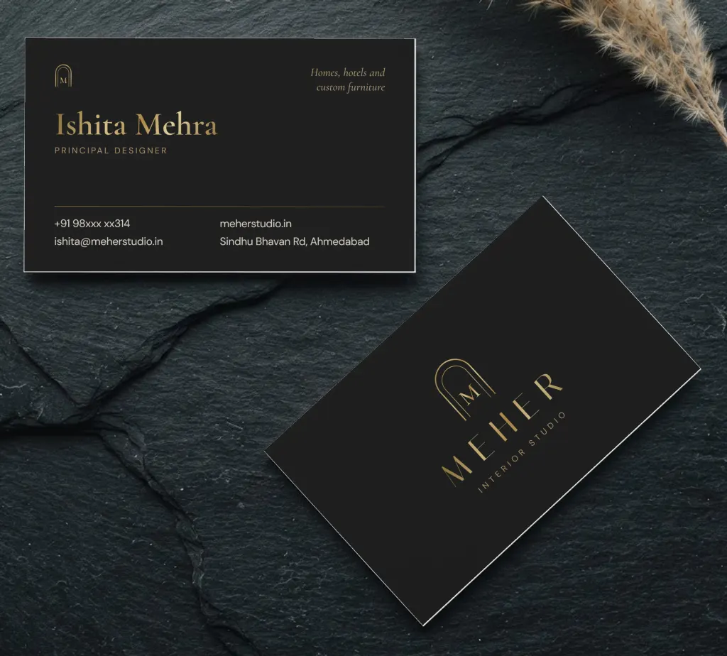 Black business cards for an interior studio in gold foil, the front with an arched M logo and the back with the designer and studio contact details, on dark stone